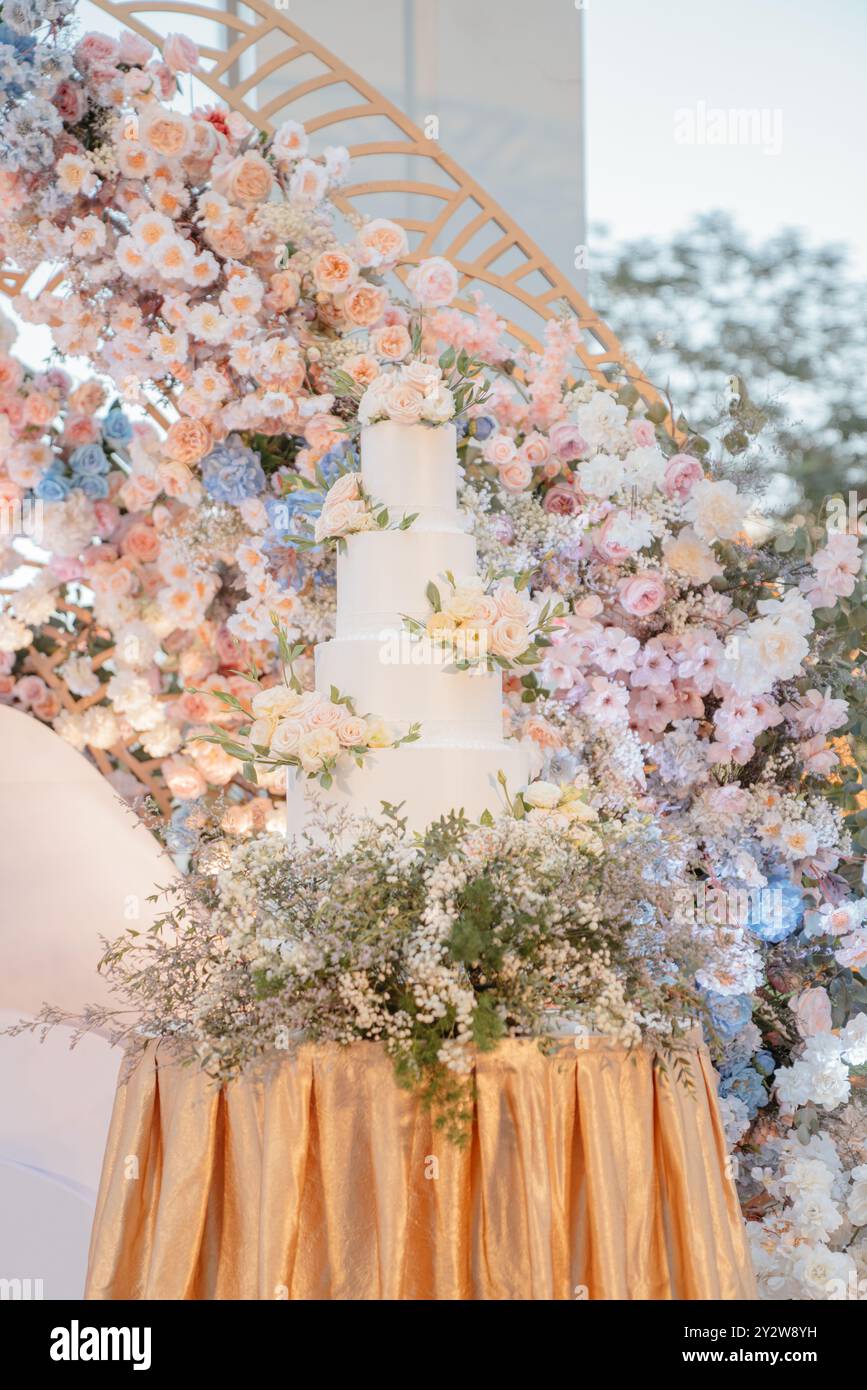 Eine wunderschön dekorierte Hochzeitstorte mit mehreren Ebenen, umgeben von einem Blumenarrangement und auf einer goldenen Tischdecke. Stockfoto