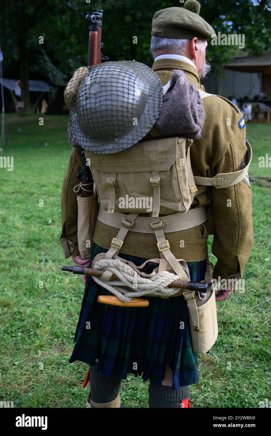 Soldat en tenue militaire écossaise, de dos, avec un sac à dos, un fusil et un casque, sur un Fond herbeux Stockfoto