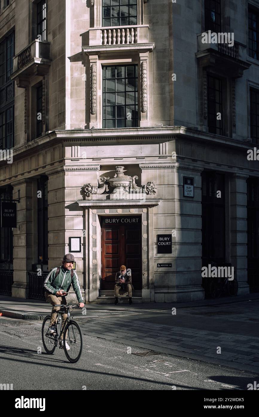 Ein Radfahrer, der am Bury Court Gebäude in London vorbeifährt, mit einer Person, die auf der Treppe sitzt Stockfoto