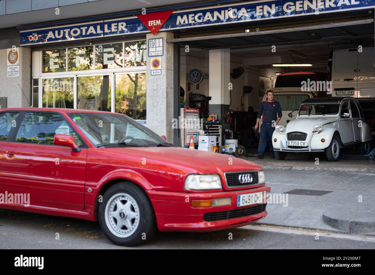 Ein Mechaniker aus einer Werkstatt in Barcelona. Im Inneren befindet sich ein alter, klassischer weißer Citroen 2cv und draußen ein schönes rotes Audi Coupé Stockfoto