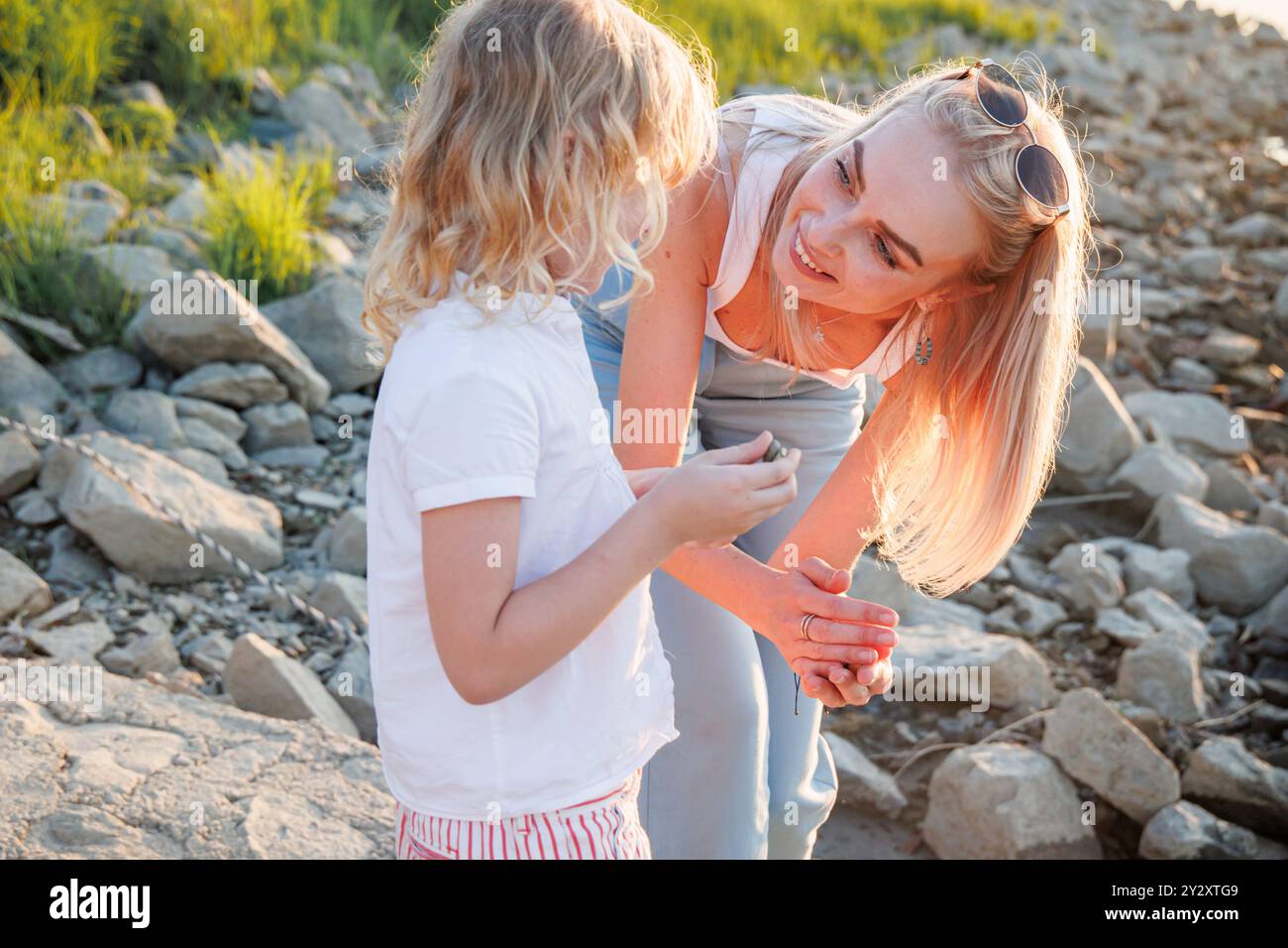 An einem angenehmen Sommerabend in Osijek, Kroatien, teilt eine Frau einen unbeschwerten Moment mit einem Kind am Fluss, umgeben von Steinen und Grün Stockfoto