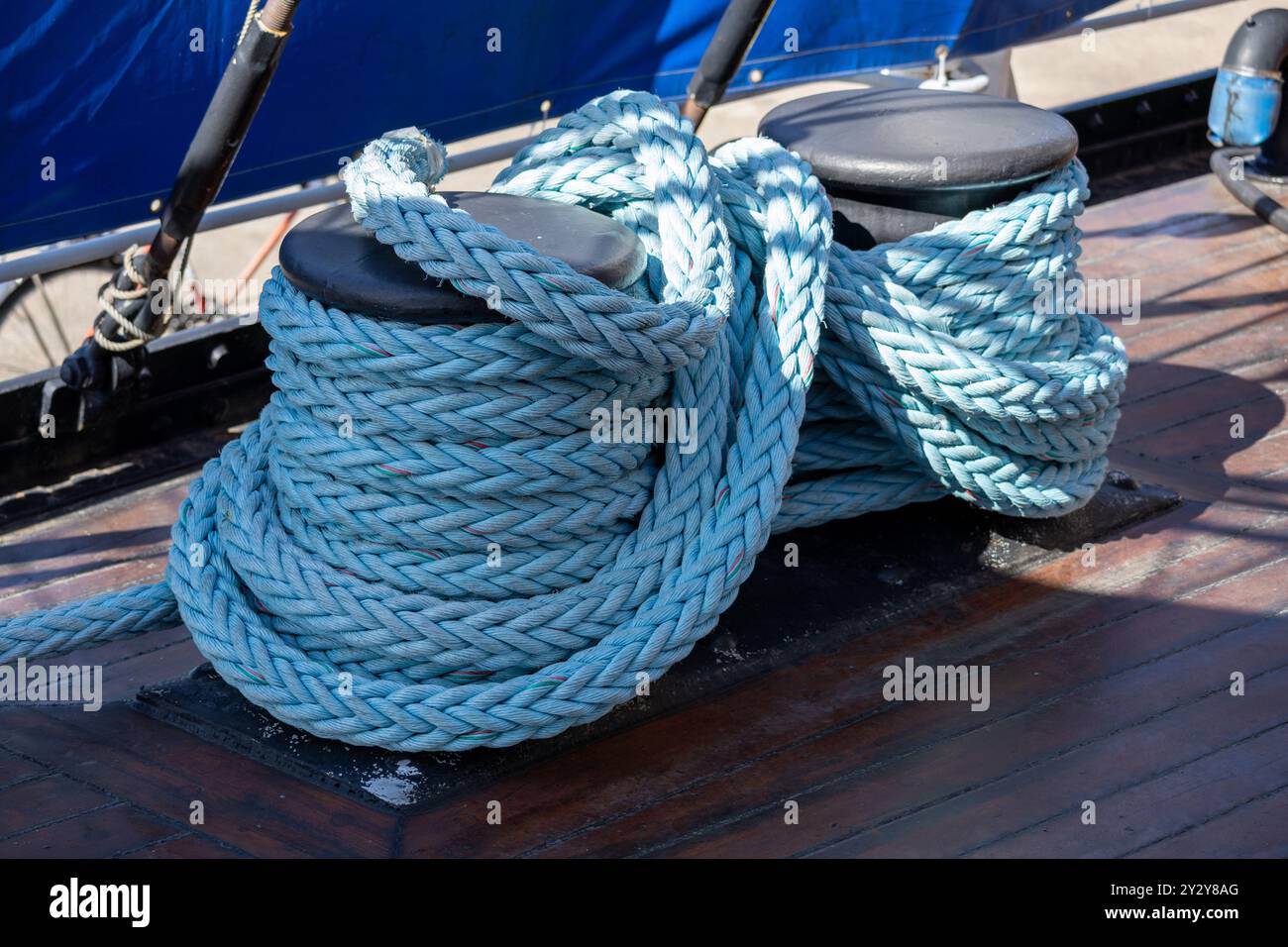 Nahaufnahme eines dicken blauen Seeseils, das auf einem Bootsdeck auf zwei schwarzen Stollen aufliegt. Der Hintergrund zeigt ein blaues Segel und ein hölzernes Deck, hochglänzend Stockfoto