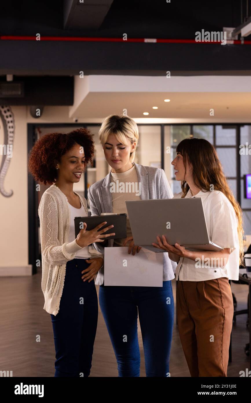 Im Büro arbeiten drei Frauen mit verschiedenen Rassen zusammen, die über Projekte mit Laptop und Tablet diskutieren Stockfoto