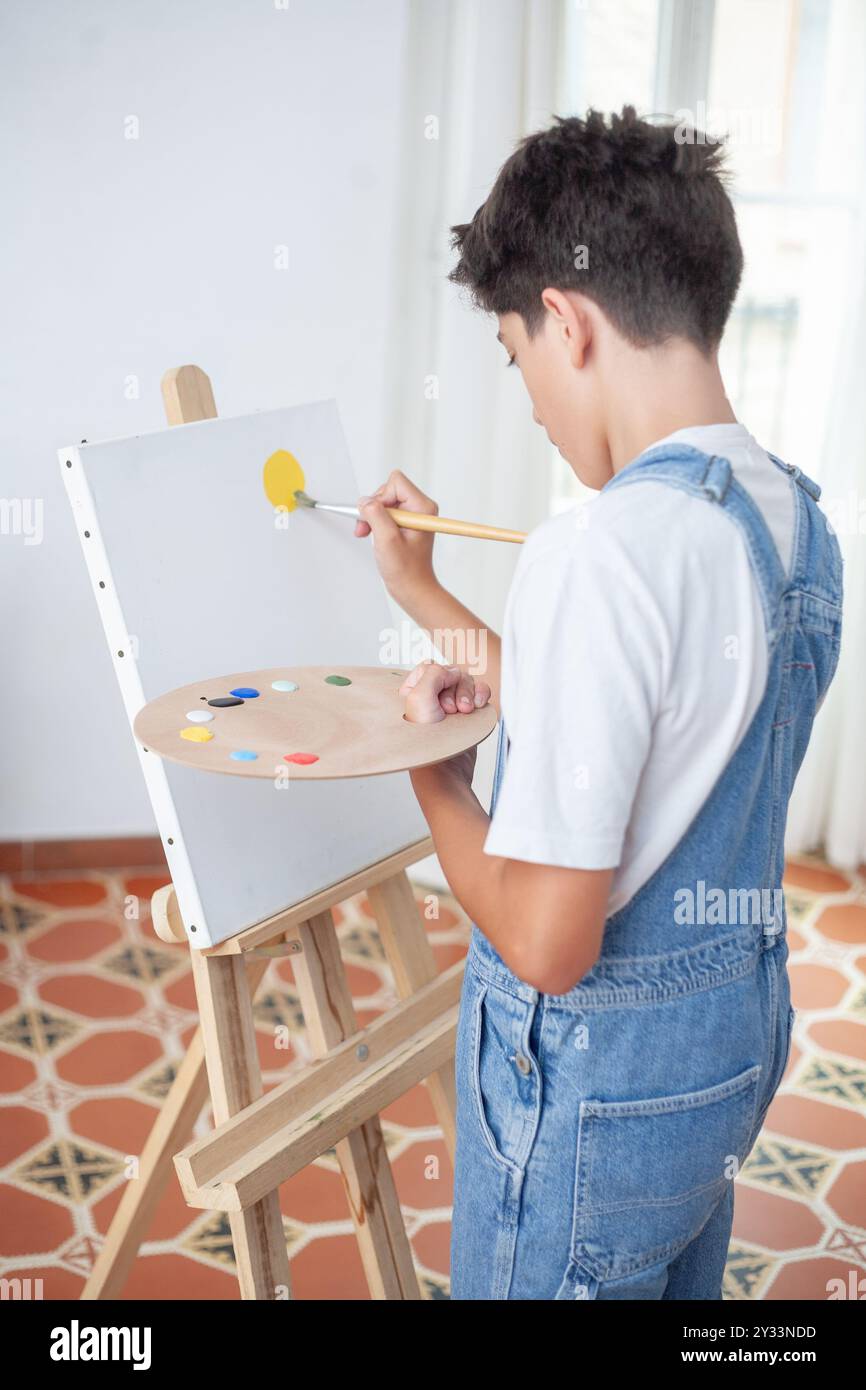 Ein kleiner Junge malt ein Bild auf eine weiße Leinwand. Er verwendet einen Pinsel und eine Farbpalette. Der Junge trägt einen blauen Jumpsuit und genießt es Stockfoto