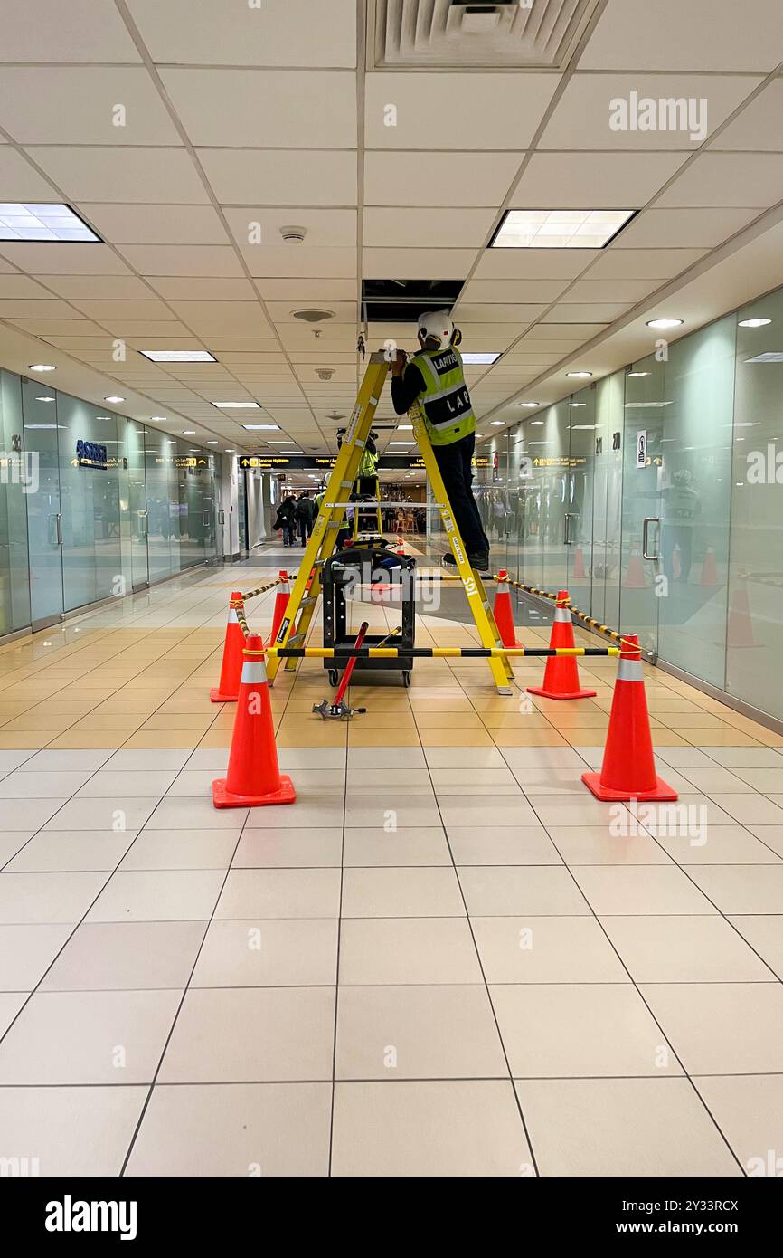 Ein Mann in einer gelben Weste steht auf einer Leiter im Flur. Es gibt orangefarbene Kegel auf dem Boden, Jorge Chavez International Airport Stockfoto