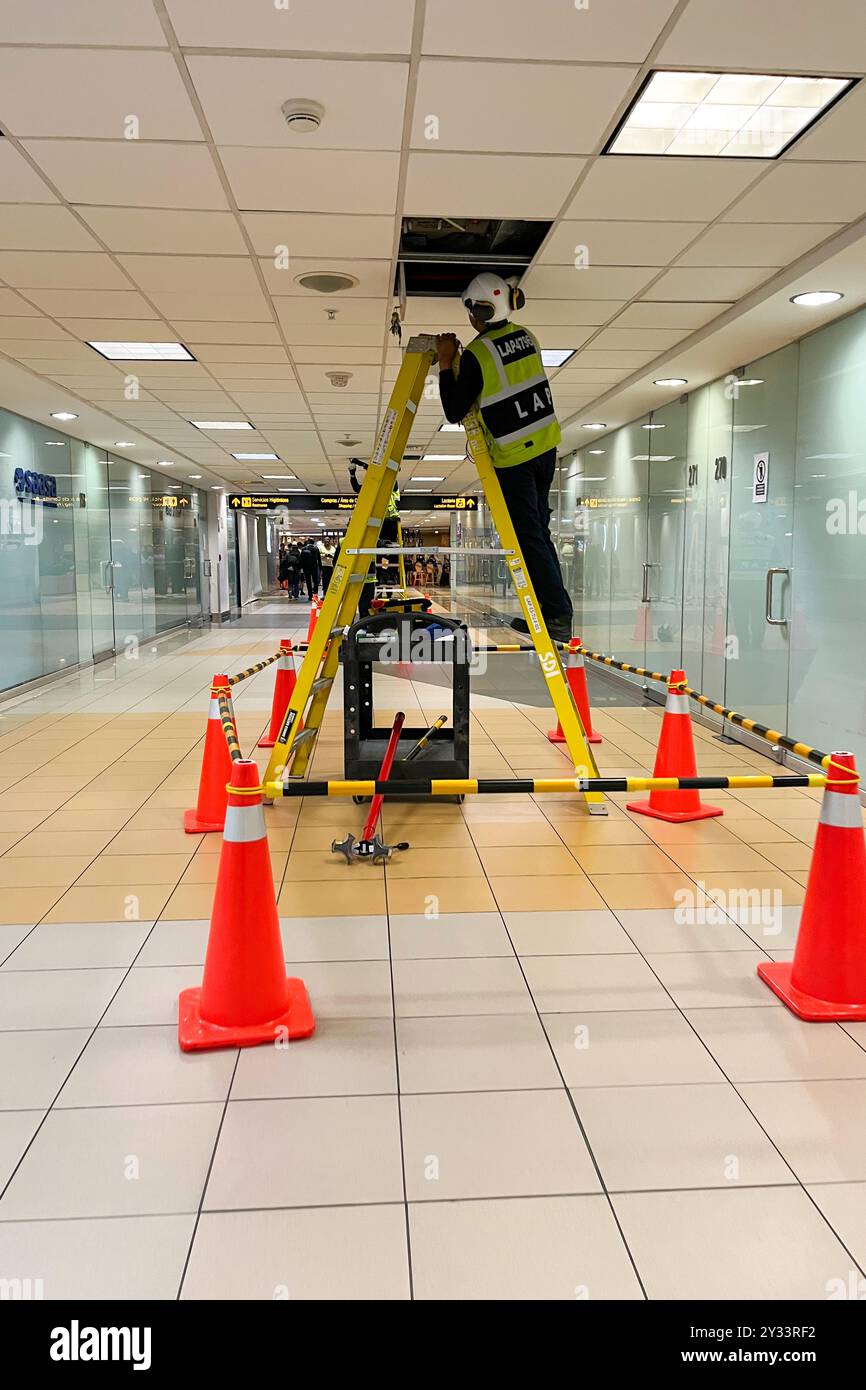 Ein Mann in einer gelben Weste steht auf einer Leiter im Flur. Im Hintergrund befinden sich orangefarbene Kegel. Jorge Chavez International Airport Stockfoto