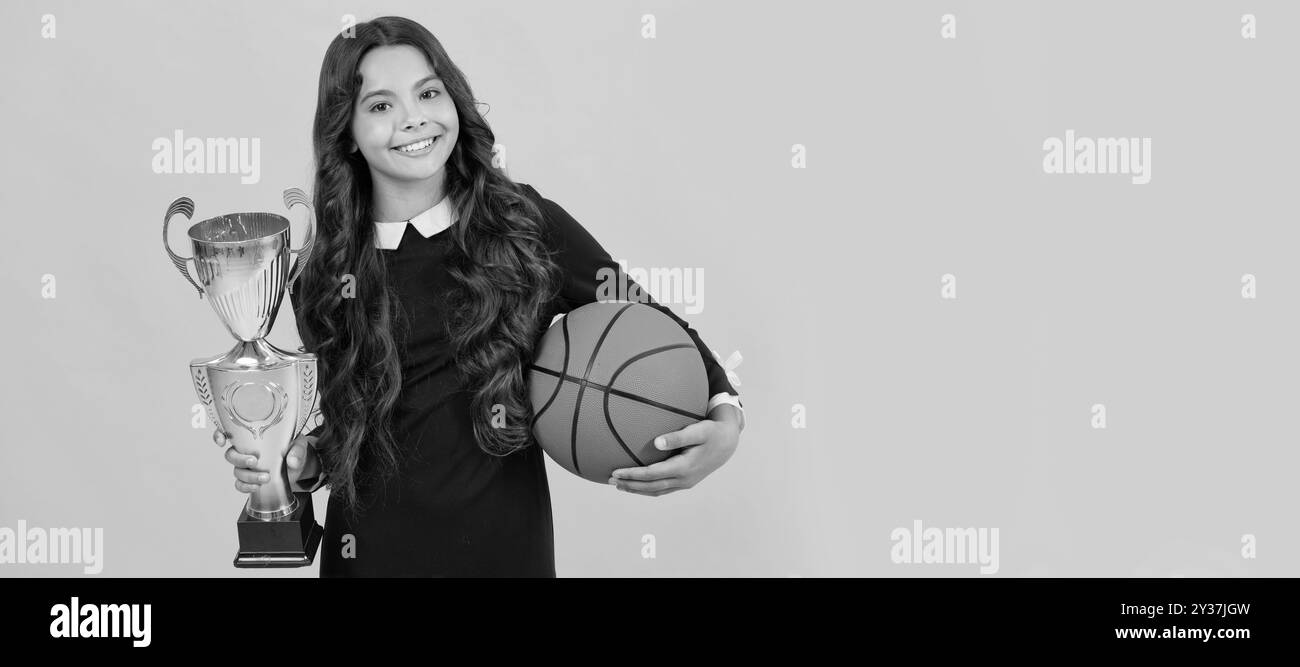 Happy teen Mädchen halten Basketball und Champion Cup auf gelbem Hintergrund, Champion. Horizontales Poster mit isoliertem Kindergesicht, Banner-Header, Kopie Stockfoto