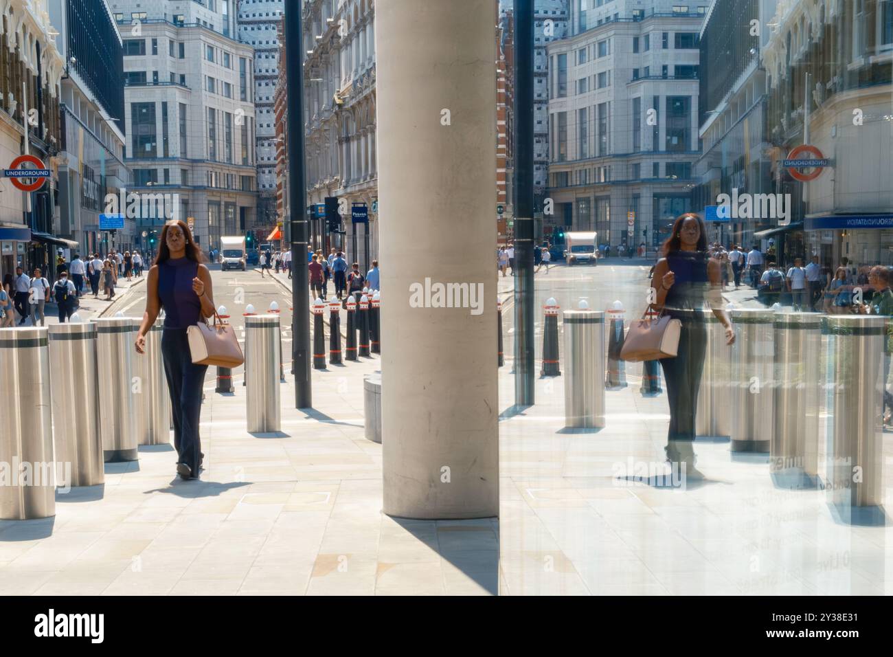 Reflexion im Fenster des weiblichen Spaziergangs in der City of London mit Stadtstraße, hohen Gebäuden, Fußgängern und festen Pollern der City of London. Stockfoto