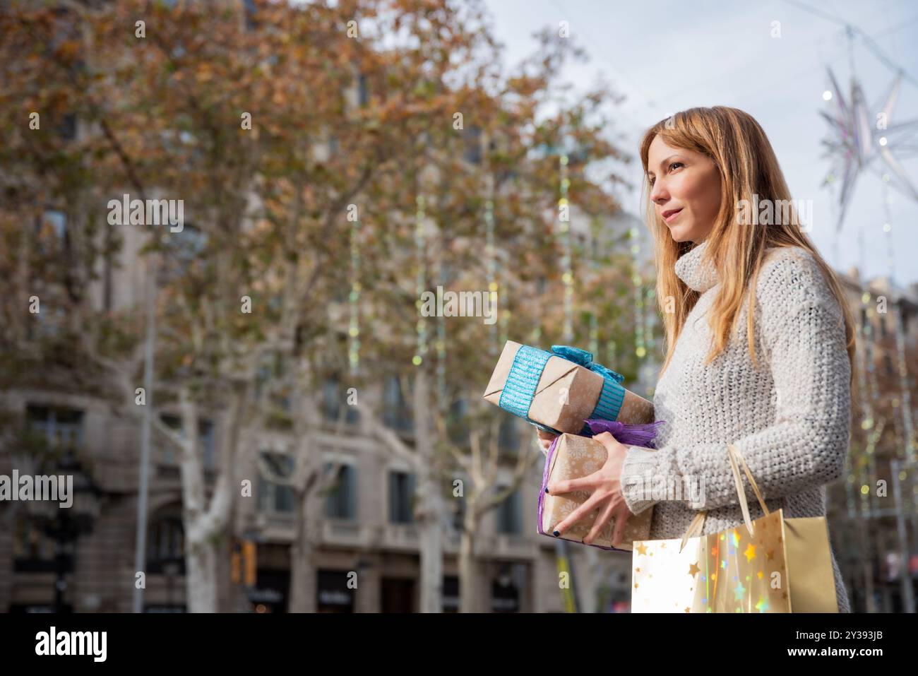 Blick von unten auf eine lächelnde Frau mit Weihnachtsgeschenken und einer goldenen Tasche auf einer festlichen Barcelona Straße, mit Platz für Text Stockfoto