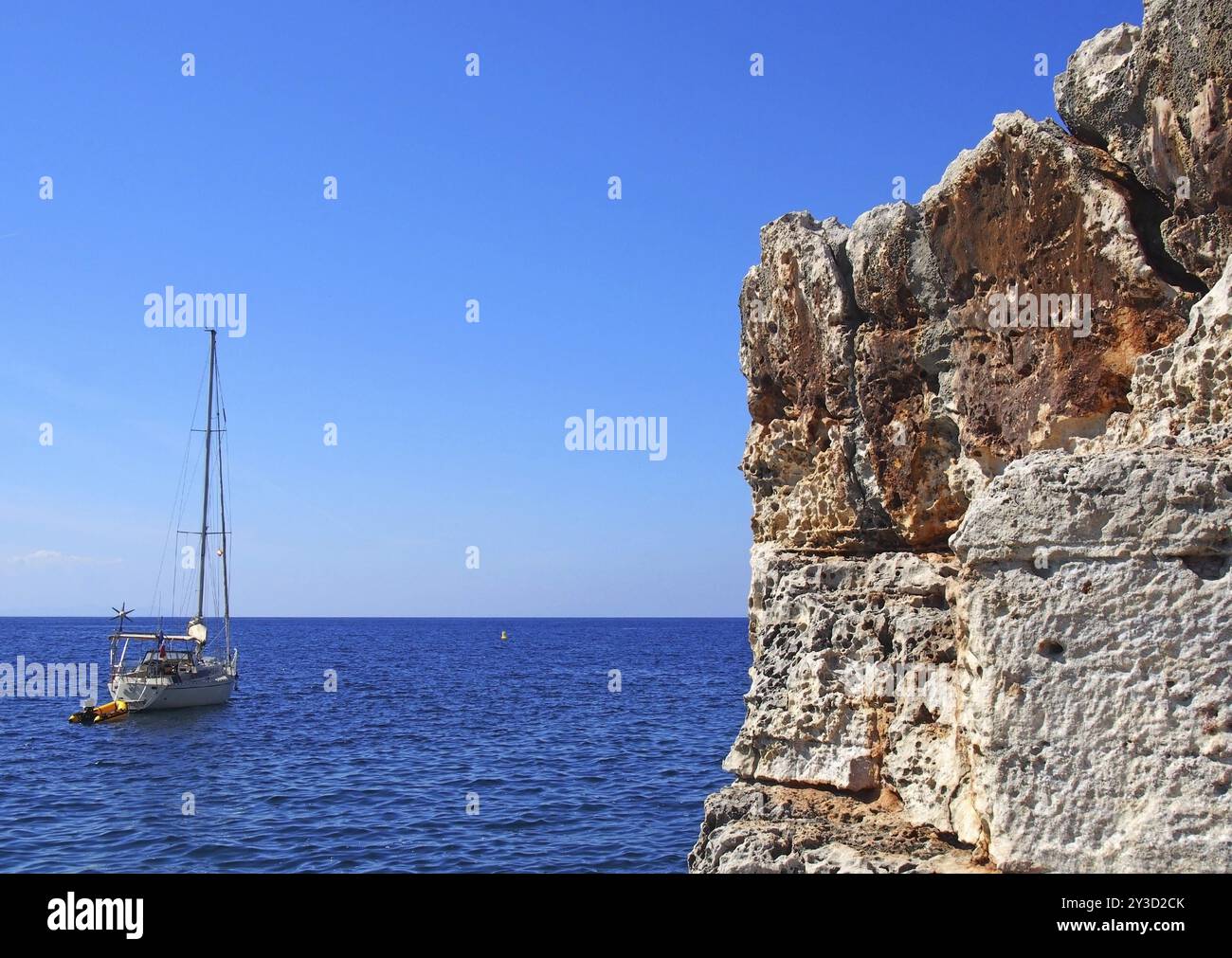 Eine Yacht auf einem hellen, ruhigen Mittelmeer neben wunderschönen zerklüfteten Klippen an der minorquinischen Küste in hellblauer Sommersonne Stockfoto