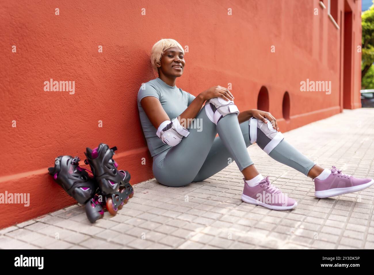 Wunderschöne afrikanische Rollschuhlaufen-Frau, die tagsüber auf dem Boden sitzt und sich auf der roten Fassade lehnt Stockfoto