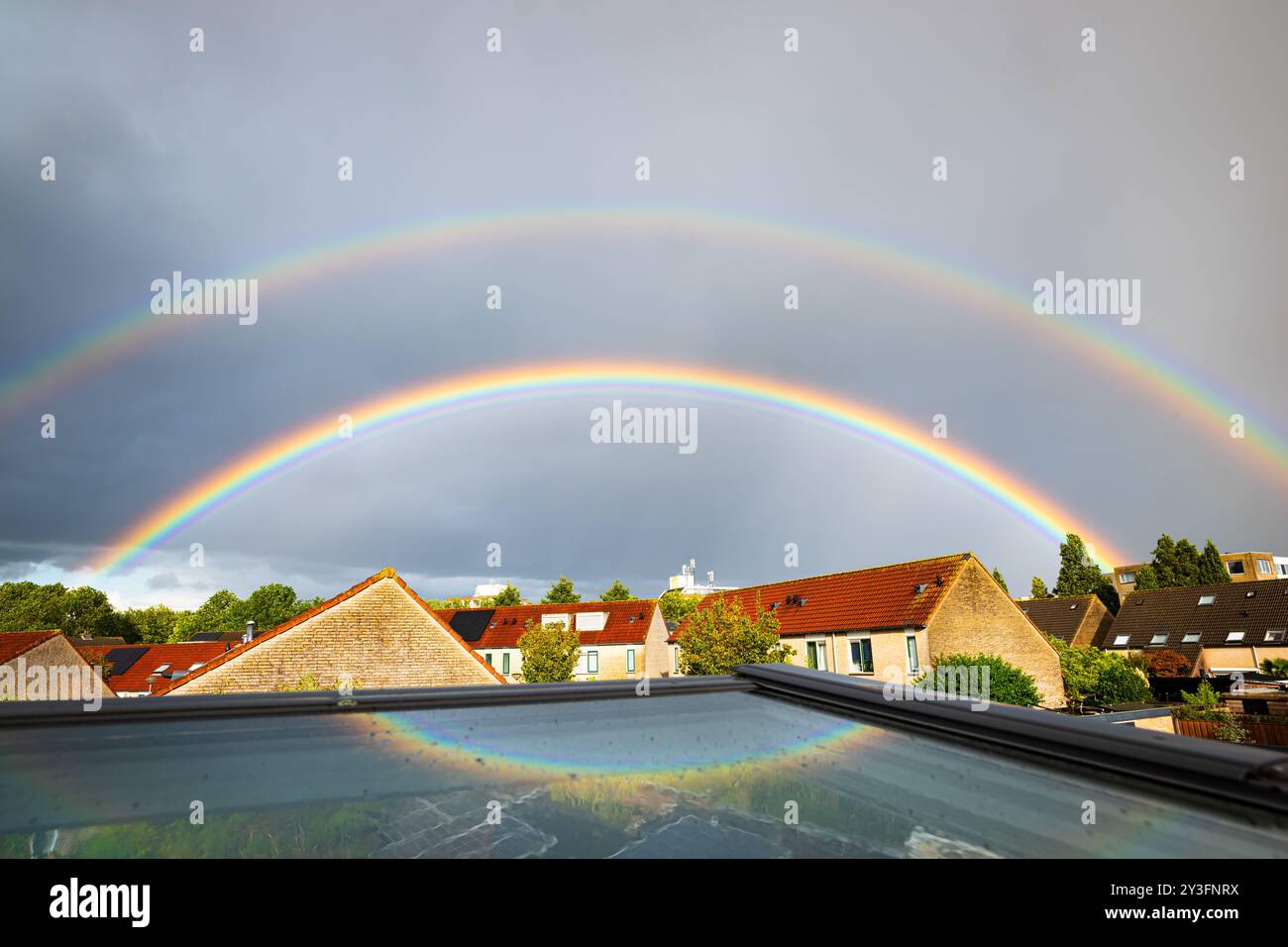 Heller bunter Doppelregenbogen über einem Wohngebiet in den Niederlanden. Ein Teil des Regenbogens wird in einem Oberlicht reflektiert. Stockfoto