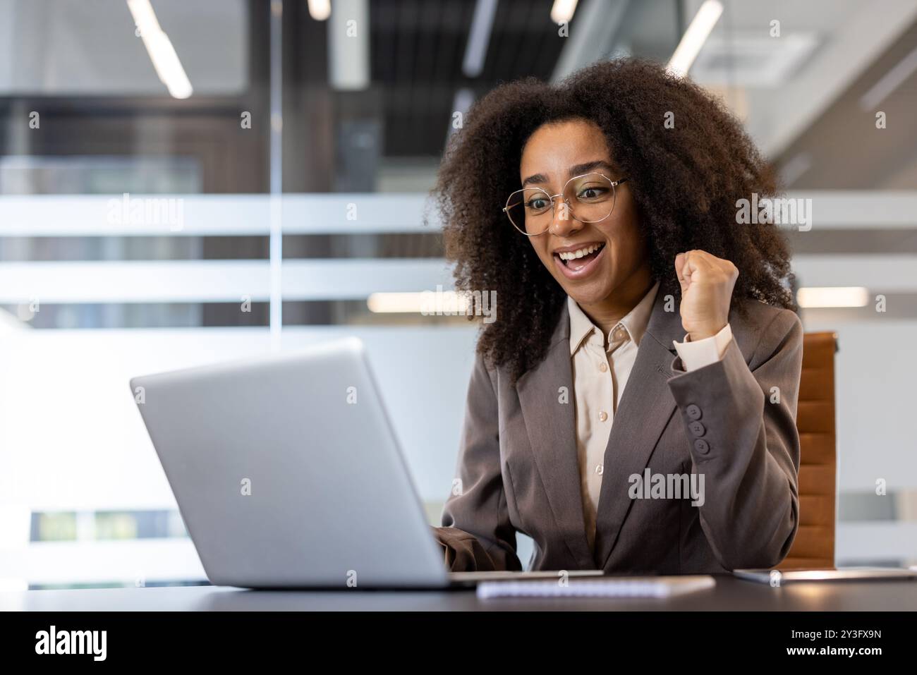 afroamerikanische Geschäftsfrau mit lockigen Haaren, die im Büro am Tisch sitzt, auf den Laptop blickt und glücklich über den Erfolg ist, mit der Hand die Siegesgeste zu zeigen und Ja zu sagen. Stockfoto
