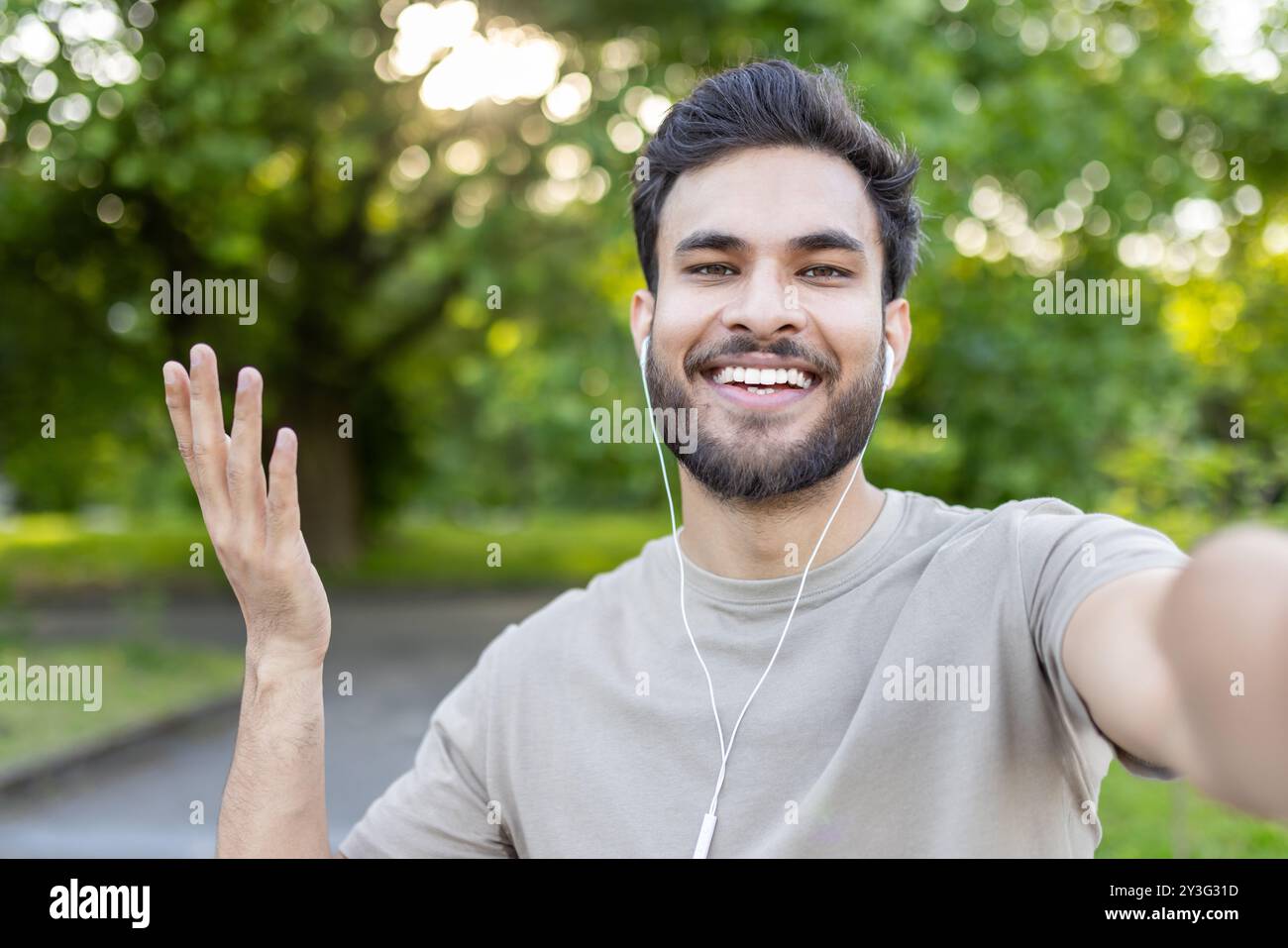 Junger Mann lächelt beim Selfie im Park mit Smartphone und Ohrhörern. Fängt Glück, Verbindung und entspannte Atmosphäre im Freien in der Freizeit ein. Stockfoto