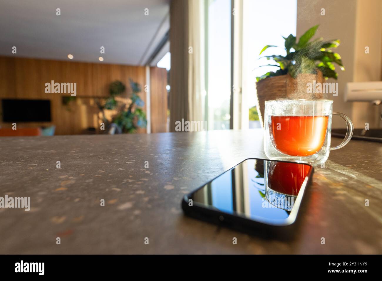 Zu Hause, Smartphone und heißer Tee auf der Küchentheke mit Sonnenlicht, das durch das Fenster strömt Stockfoto