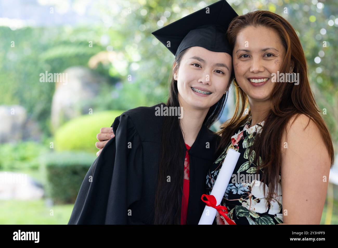 Im Garten, asiatische Tochter in Mütze und Kleid mit Diplom, posiert mit stolzer Mutter Stockfoto