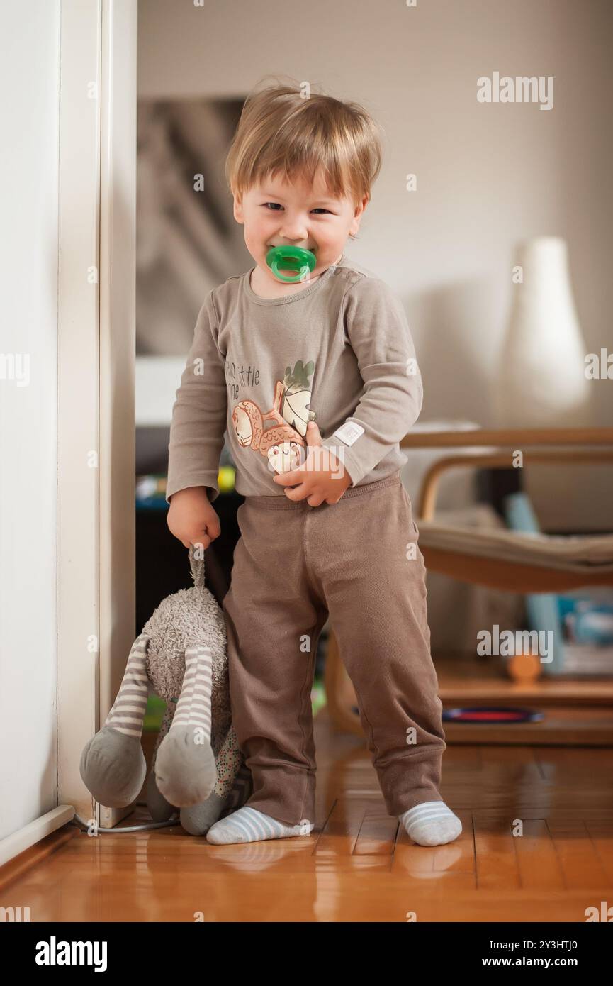 Ein fröhliches kleines Kind steht vor der Tür und hält sich an einen geliebten Plüschhasen. Die warme Innenbeleuchtung sorgt für eine gemütliche Atmosphäre und verbessert das Spiel Stockfoto