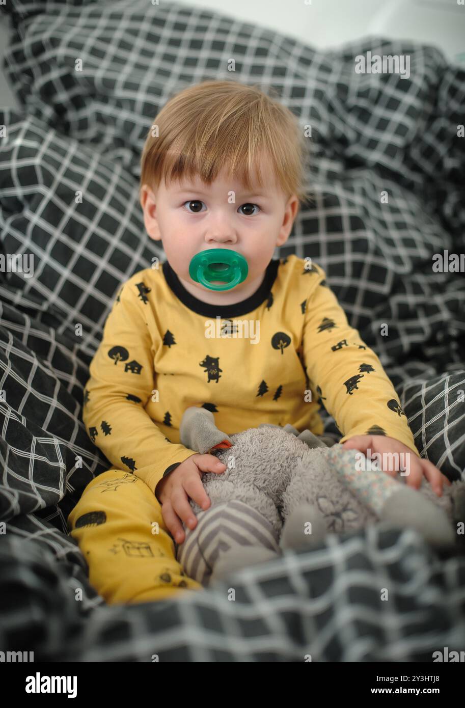 Ein fröhliches Kleinkind sitzt bequem zwischen einem Meer aus gemusterten Decken, hält ein weiches Spielzeug und trägt einen entzückenden Pyjama, der Wärme und Unschuld ausstrahlt Stockfoto