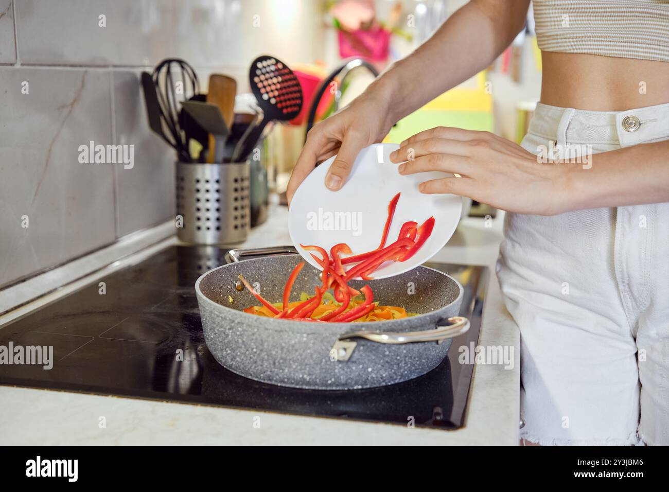 Eine Person bereitet ein Gericht zu, indem sie gehackte rote Paprika in eine Pfanne auf einem Herd gibt. Die Küche verfügt über moderne Geräte und eine Auswahl an kochutensilien Stockfoto