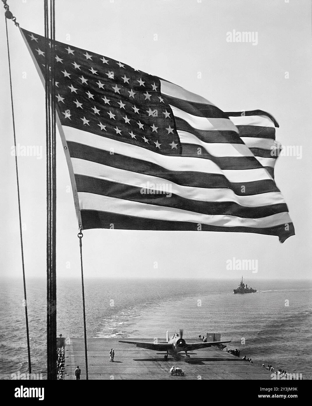 Die Stars and Stripes winken über einen Flugzeugträger der US Navy auf See. Auf diesem Flugdeck befindet sich ein Grumman 'Avenger' TBM, ein Torpedoflugzeug; östlich, das hinter dem Träger zum Hafen abbiegt, befindet sich ein Escort-Zerstörer. November 1942. Foto der US Navy. Stockfoto