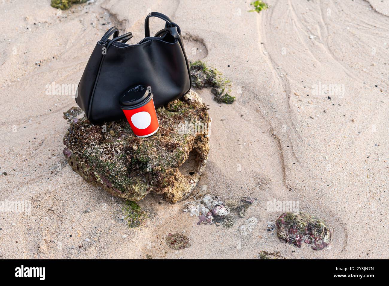 Eine stilvolle schwarze Handtasche liegt auf einer felsigen Oberfläche am Strand neben einer roten Kaffeetasse mit weißem Kreis. Der Sandstrand hat verstreute Felsen A Stockfoto