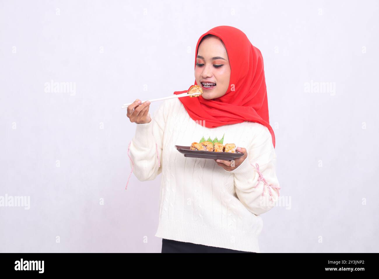 Fröhliche indonesienfrau im Hijab hält Essstäbchen an das Gericht und trägt einen Teller mit Sushi (japanisches Essen) während der Präsentation. Schöne Muslime Stockfoto