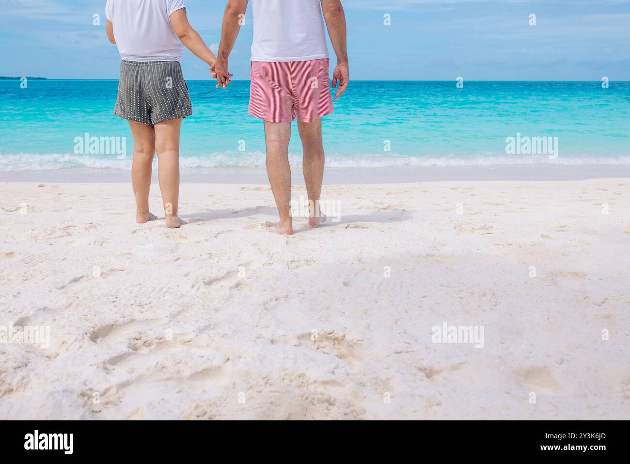 Nahaufnahme eines jungen verheirateten Paares in Shorts und T-Shirts am Meer. Mann und Frau halten Hände und genießen die Meereslandschaft. Ansicht von hinten. Ehemann und Ehefrau Stockfoto