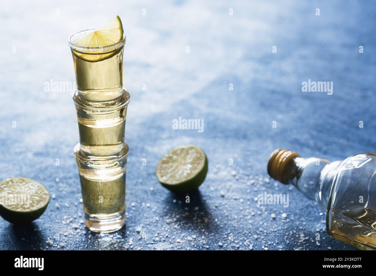 Goldener Tequila im Glas mit Salz und Limette, auf einem blauen Tisch, Hintergrundbeleuchtung. Drei Tequila-Schüsse aufeinander gestapelt. Traditionelles mexikanisches Getränk Stockfoto