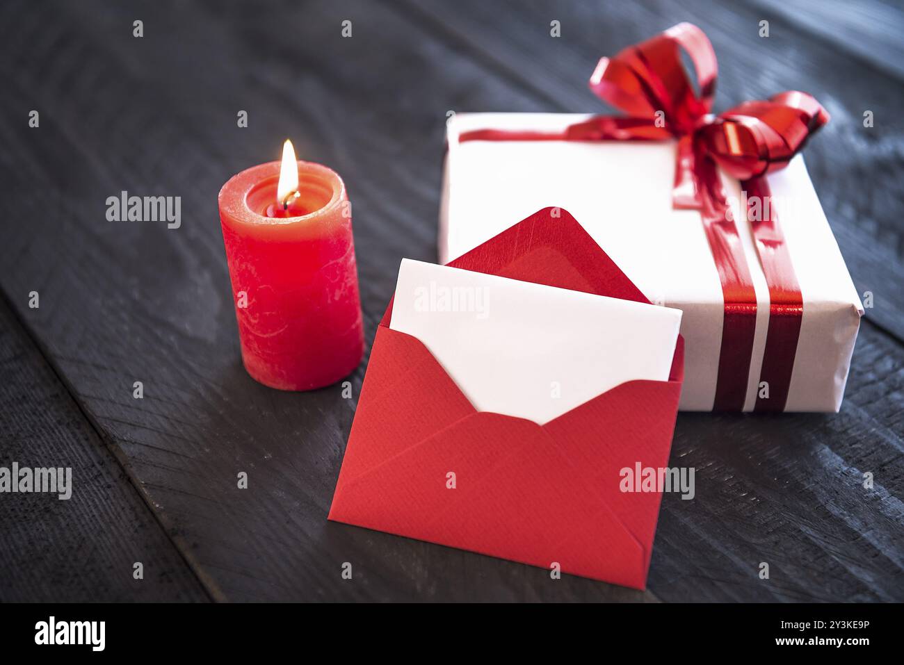 Ein roter, leerer Umschlag lehnte sich an ein stilvoll gewickeltes Geschenk, gebunden mit rotem Band und Schleife, neben einer zündeten Kerze auf einem alten Holztisch Stockfoto