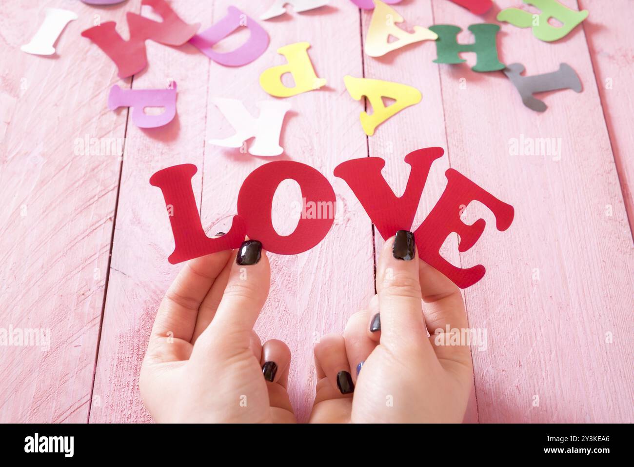 Frauenhände mit schwarzen Nägeln, die das Wort Liebe aus roten Papierbuchstaben über einem rosafarbenen Holztisch halten, mit bunten Buchstaben im Hintergrund Stockfoto