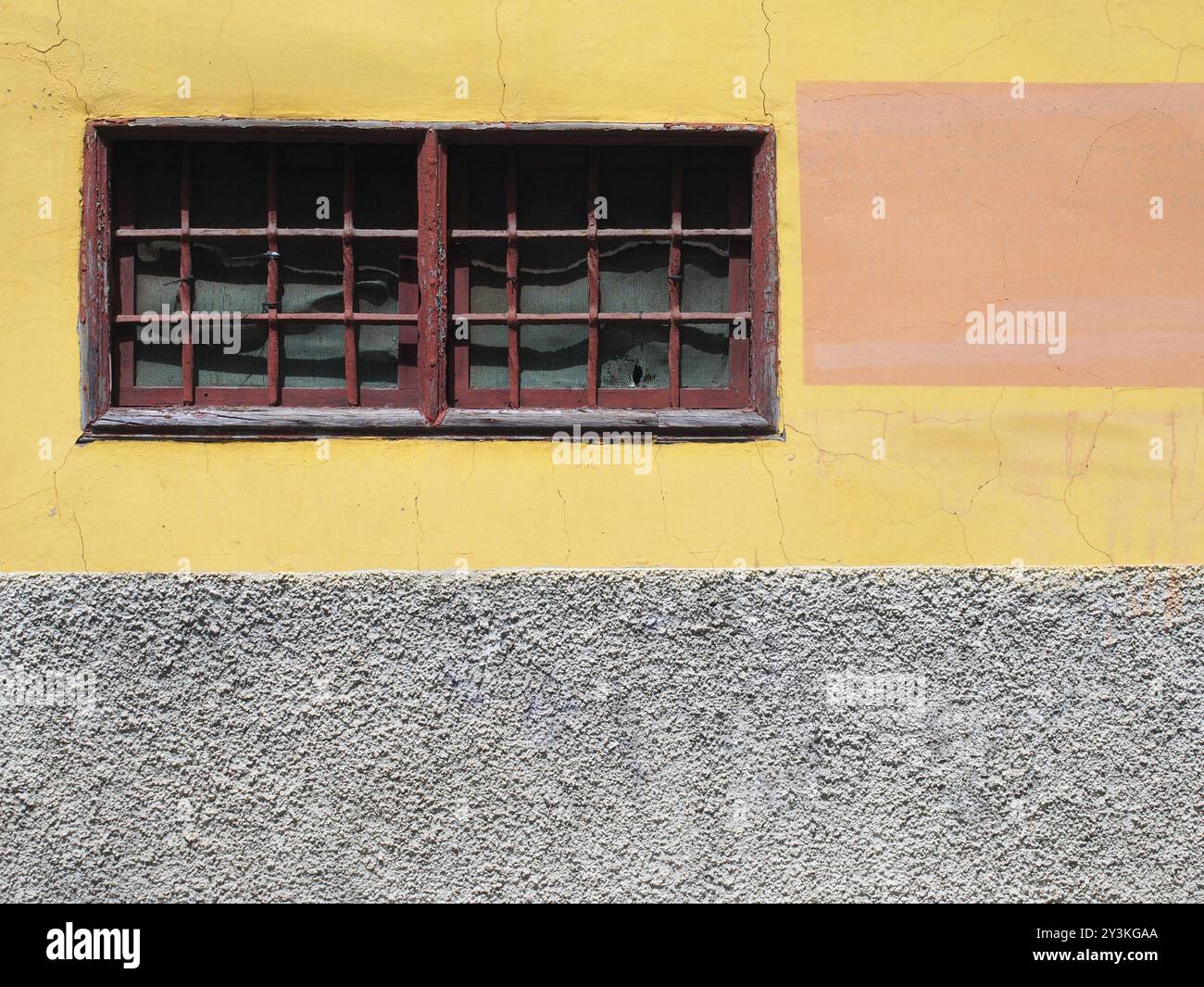 Alte, gelb-graue Betonwand mit schmalen, rot gerahmten Fenstern mit Gittern und einer stark rissigen Textur Stockfoto