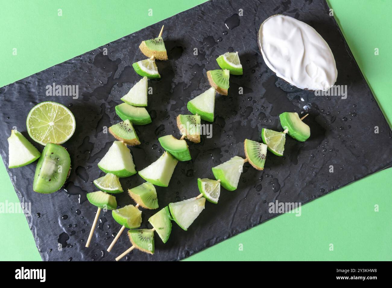 Grüne Tischplatte mit schwarzem Schneidebrett mit Spießen aus grünen Früchten und Joghurtsauce. Vegetarische Mahlzeit. Konzept für gesunde Ernährung. Detox Food Stockfoto