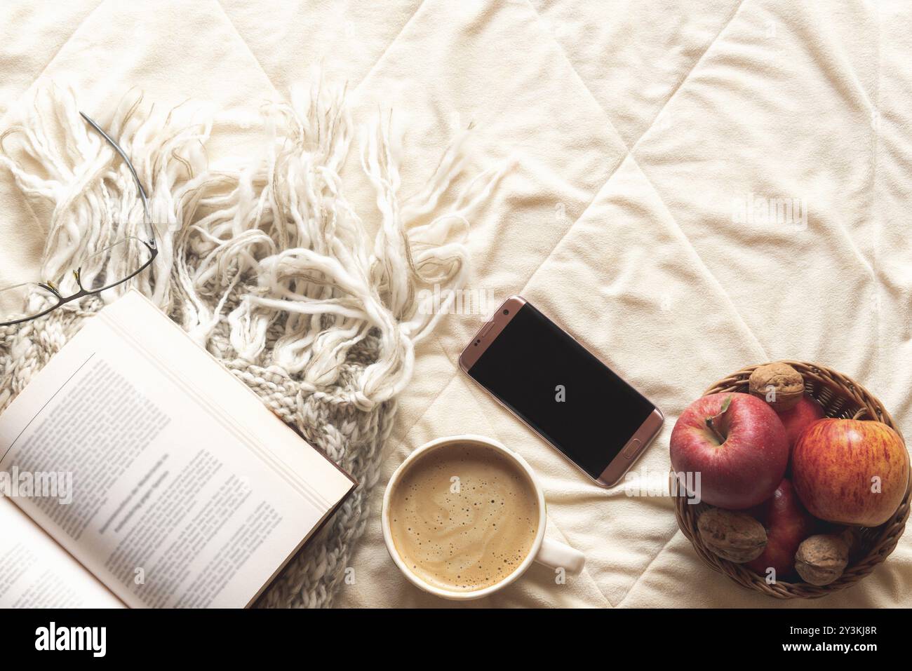 Gemütliches Konzept mit offenem Buch auf einem Wolltuch, einem Korb mit Äpfeln und Nüssen, einem Telefon und einer Tasse heißem Kakao auf einer flauschigen Fleecedecke Stockfoto