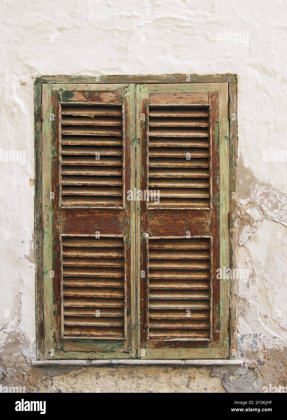 Alte geschlossene, grün lackierte, braune Fensterläden in einer weißen Wand mit zerrissenem Zement und Putz Stockfoto