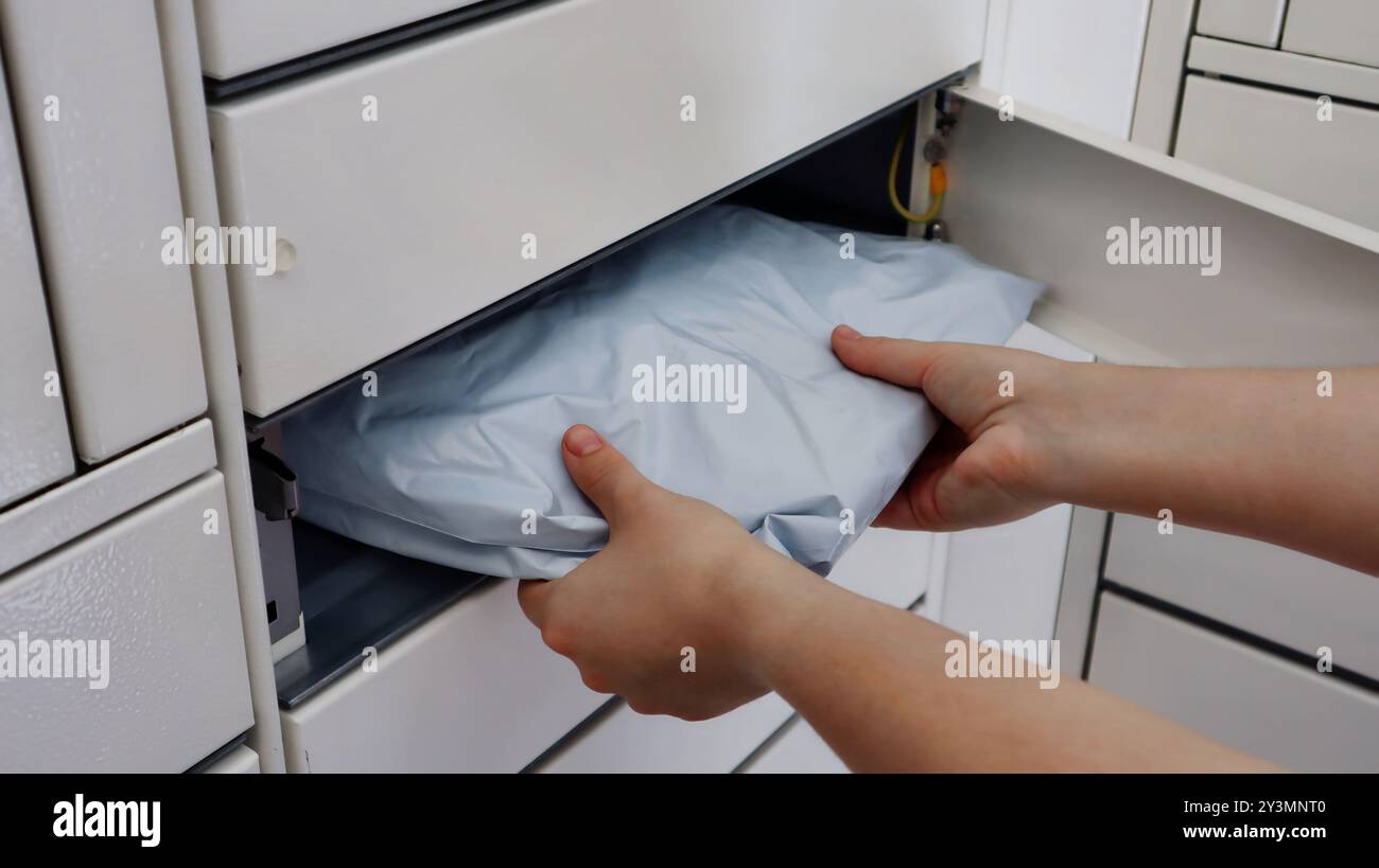 Eine Person, die eine versiegelte Verpackung in ein weißes Fach oder ein Ablagefach legt. Die Hände schieben das graue Paket vorsichtig in den offenen Schlitz. Stockfoto