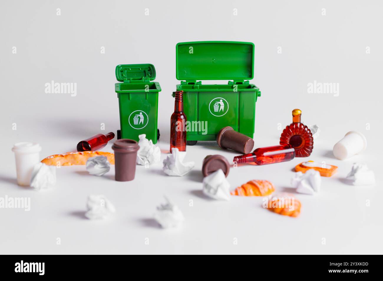 Miniatur-Aufbau mit verschiedenen Abfällen wie Lebensmitteln, Plastikbechern und Glasflaschen, die von kleinen grünen Recyclingbehältern auf neutralem Hintergrund verstreut sind. Stockfoto