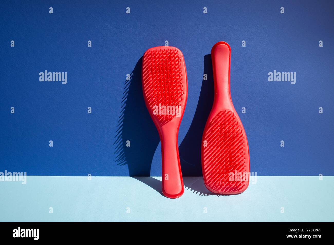 Aufrecht stehende rote Haarbürste an der Wand, beleuchtet durch Tageslicht, wirft definierten Schatten. Die dynamische Kombination in Rot und Blau sorgt für zeitgemäßes Flair und eine visuelle Komposition Stockfoto