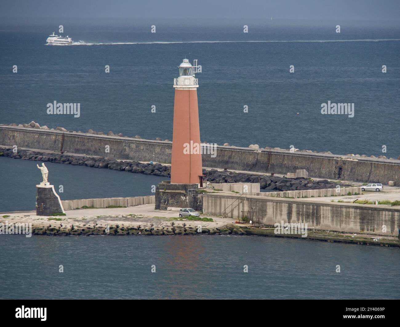 Leuchtturm am Hafen mit vorbeifahrendem Boot und Schiff am Horizont, bewölkter Himmel, neapel, mittelmeer, italien Stockfoto