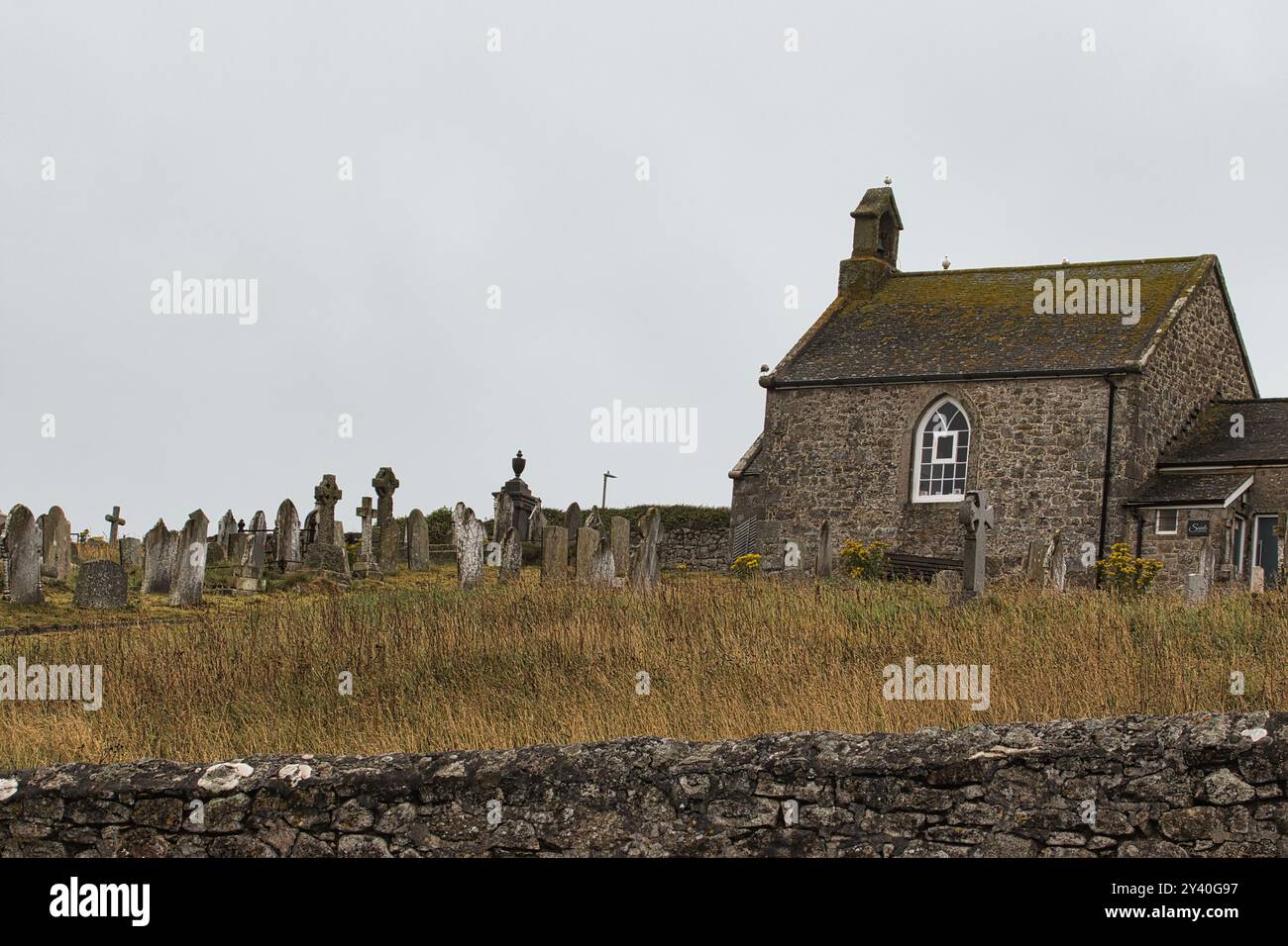 Eine historische Steinkirche neben einem Friedhof mit verwitterten Grabsteinen und einem grasbewachsenen Hügel. Der Himmel ist bedeckt und schafft eine düstere Atmosphäre. Stockfoto