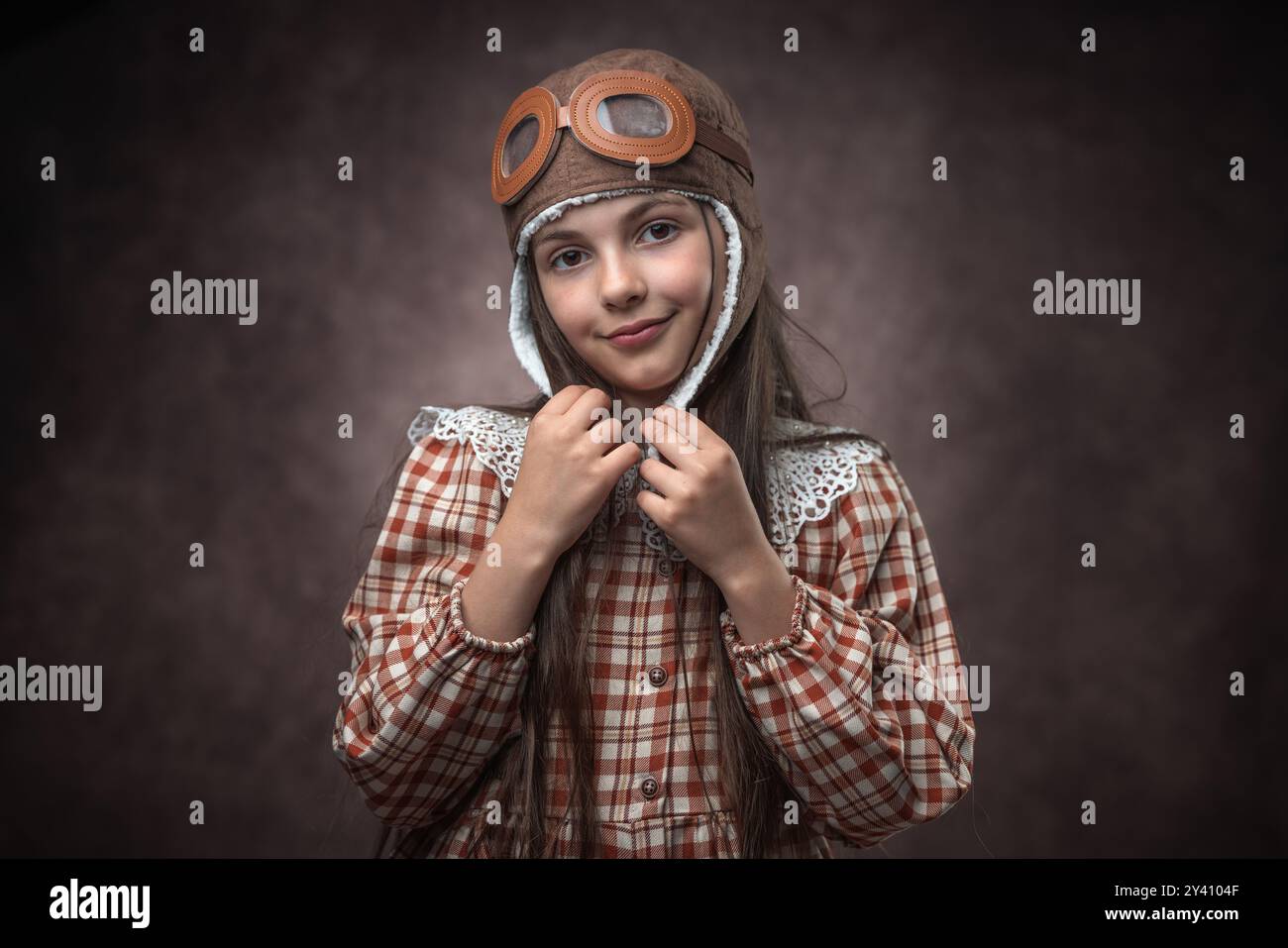 Retro-Porträt eines kaukasischen Mädchens in Vintage-Kleidung mit Fliegerhut vor einem alten braunen Tapetenhintergrund Stockfoto