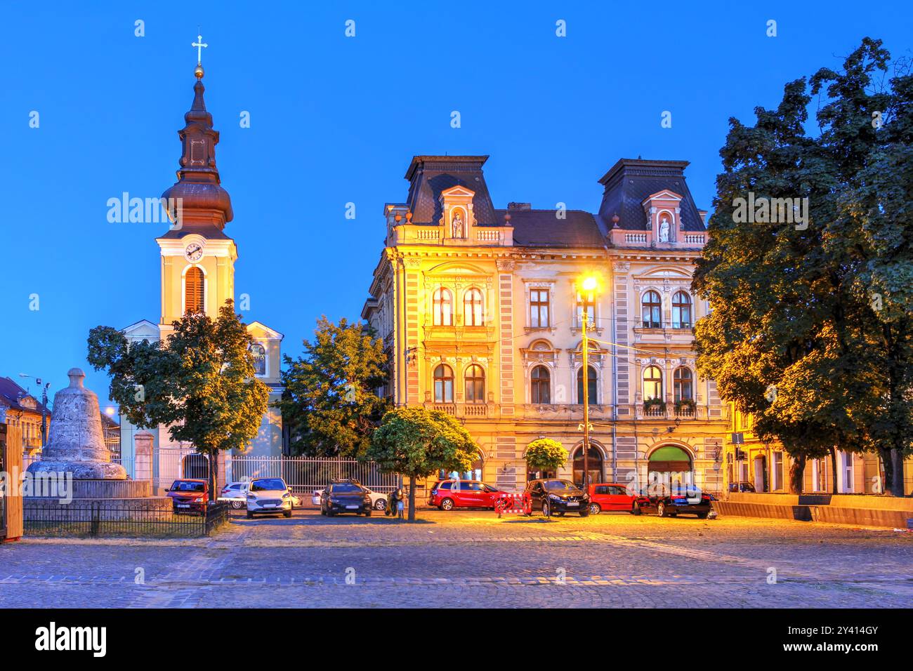 Nacht in Piata Traian (Traian-Platz) in Timisoara, Rumänien mit der St. Georgskirche und dem Palast der serbischen Gemeinde. Stockfoto