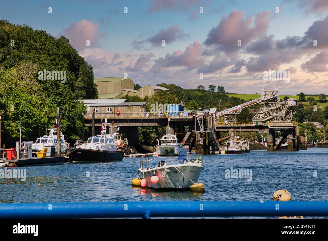 Ein malerischer Blick auf einen Hafen mit Booten im Vordergrund. Im Hintergrund befindet sich ein Dock mit Industriegebäuden und üppigem Grün. Der Himmel ist Stockfoto