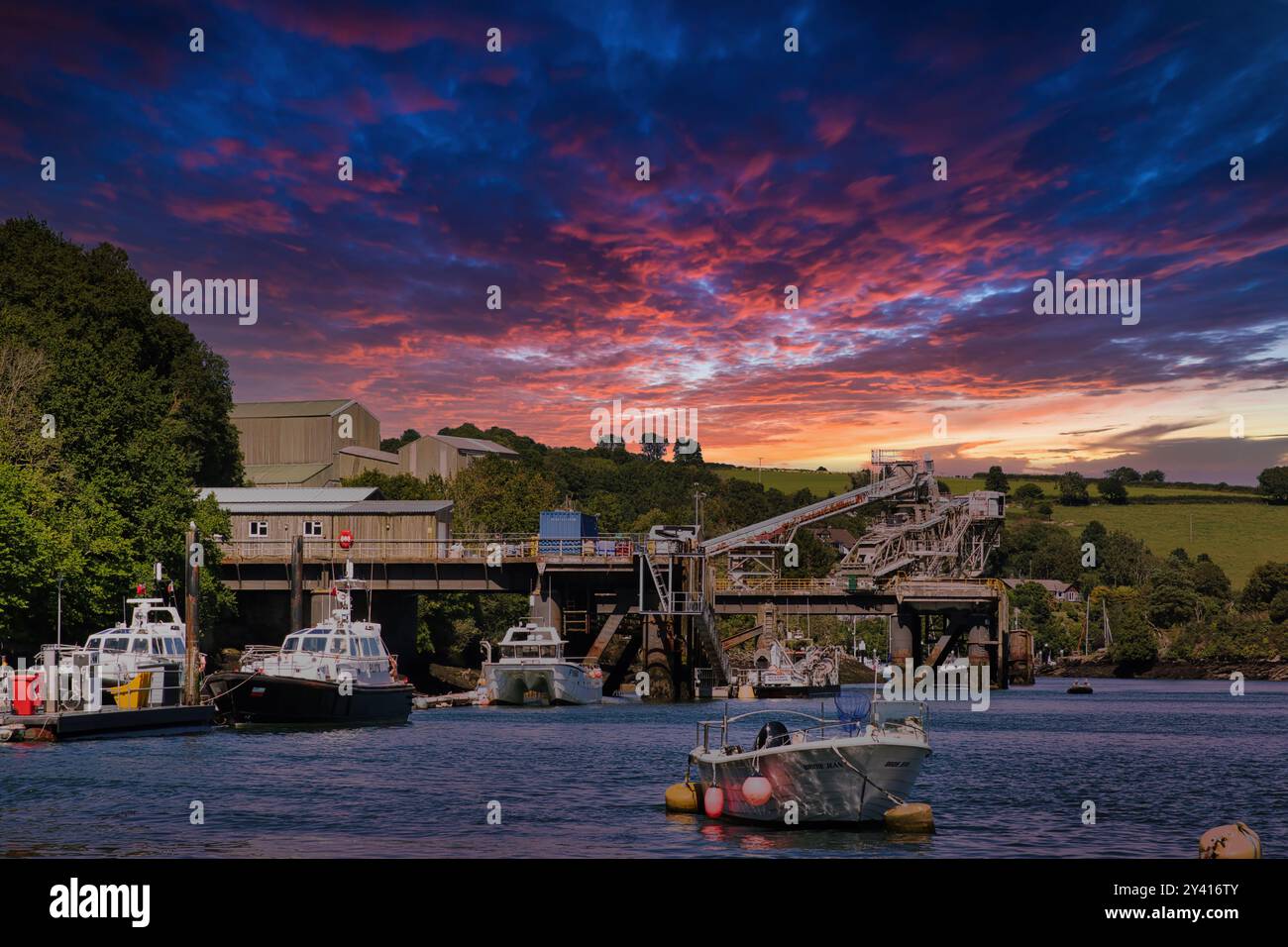 Ein malerischer Blick auf einen Hafen bei Sonnenuntergang, mit Booten im Vordergrund und einem Dock mit Industriebauten im Hintergrund. Der Himmel ist voller Stockfoto