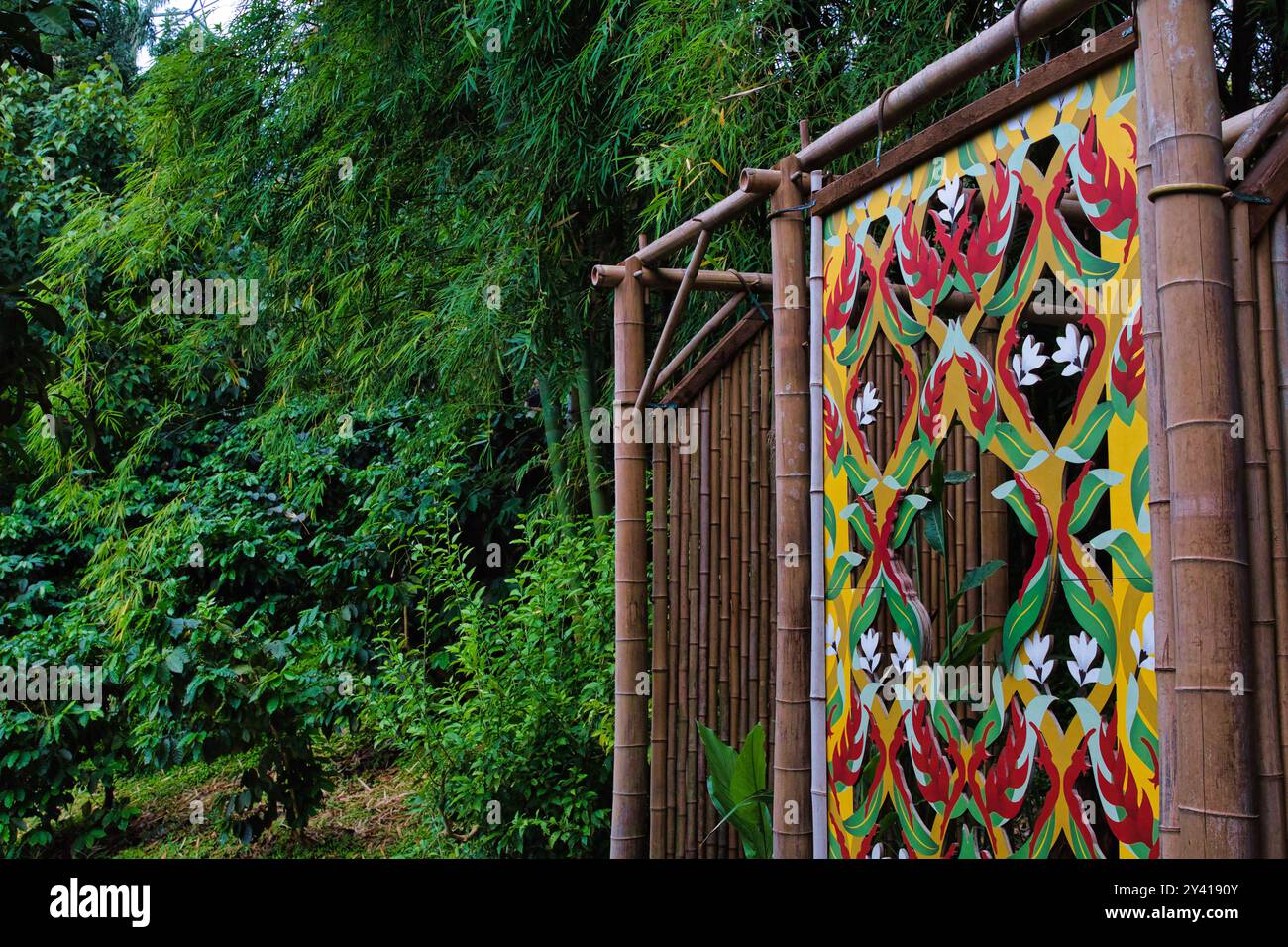 Ein lebendiges, farbenfrohes Wandbild auf einem Bambuszaun, umgeben von üppigem Grün. Das Wandbild zeigt komplizierte Blumenmuster in Rot, Gelb und Grün Stockfoto