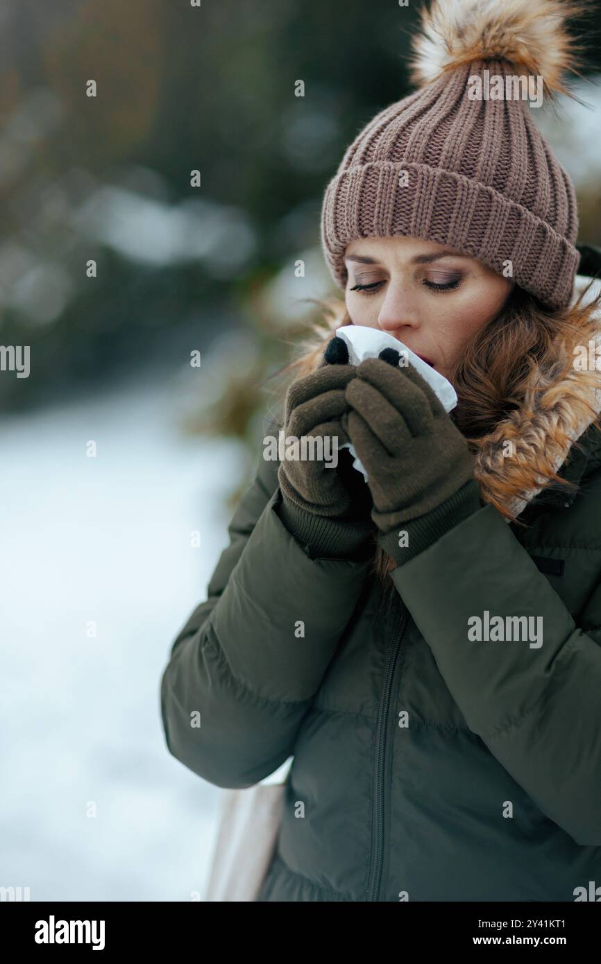 Traurige, moderne Frau mittleren Alters im grünen Mantel und braunem Hut draußen im Stadtpark im Winter mit Fäustlingen, Servietten-Blasnase und Beanie-Hut. Stockfoto