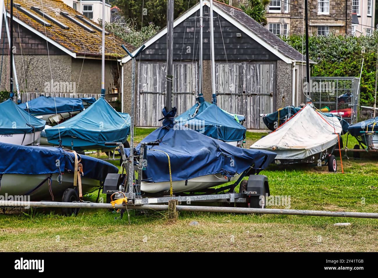 Eine Ansammlung von Booten, die mit blauen Planen bedeckt sind und auf einem grasbewachsenen Gelände in der Nähe eines Holzschuppen geparkt sind. Die Szene fängt eine ruhige maritime Atmosphäre mit einem Hi ein Stockfoto
