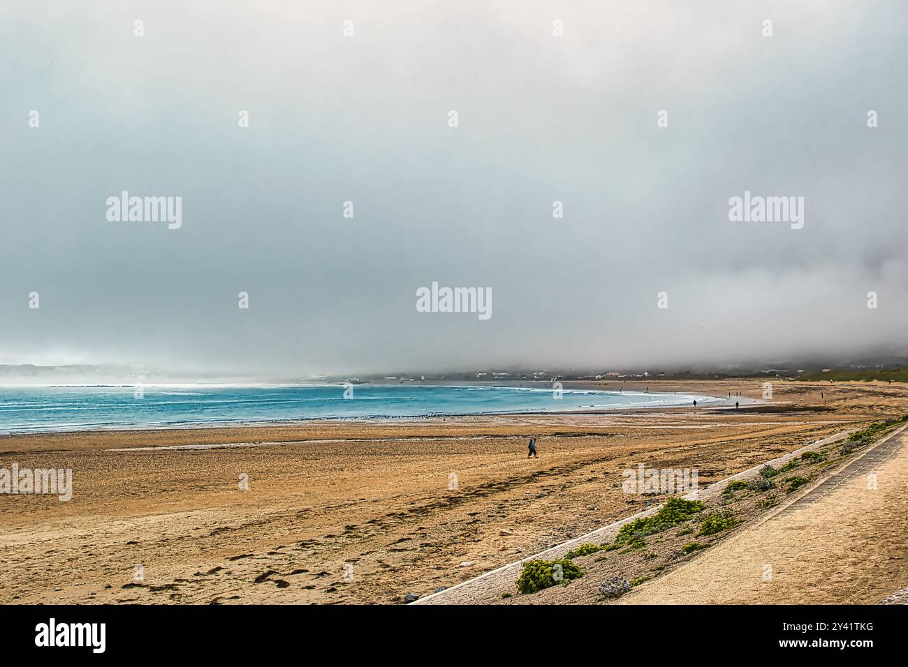 Eine ruhige Strandszene mit einem breiten Sandstrand und sanften Wellen. Der Himmel ist bedeckt mit Nebel, was eine stimmungsvolle Atmosphäre schafft. Ein paar Leute können gesehen werden Stockfoto