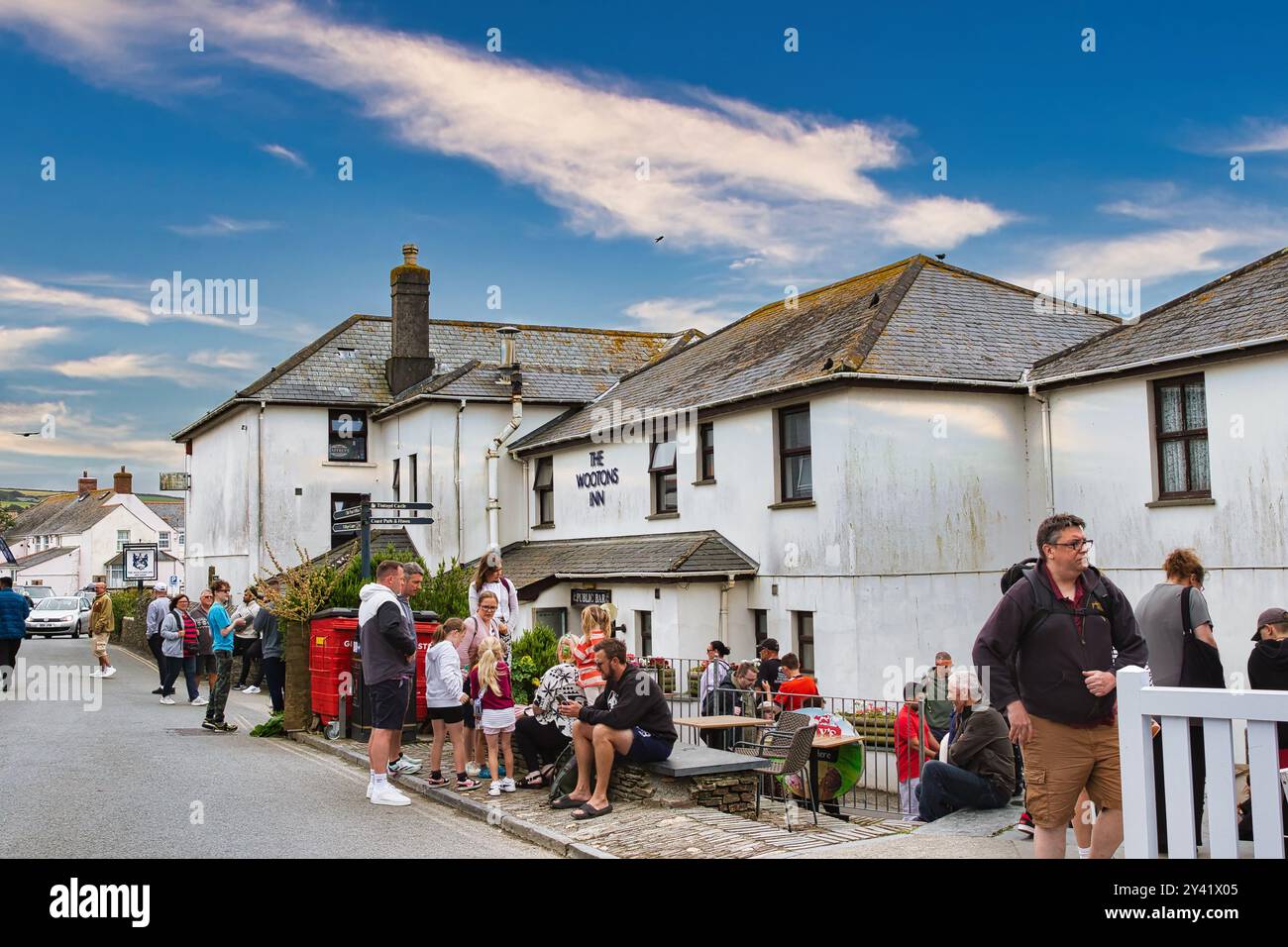 Eine lebhafte Szene vor einem Pub mit Leuten, die sich auf der Straße und auf der Terrasse versammelten. Das Gebäude ist weiß mit einem Schild mit der Aufschrift "The Wooding Inn". Der Himmel ist blau Stockfoto