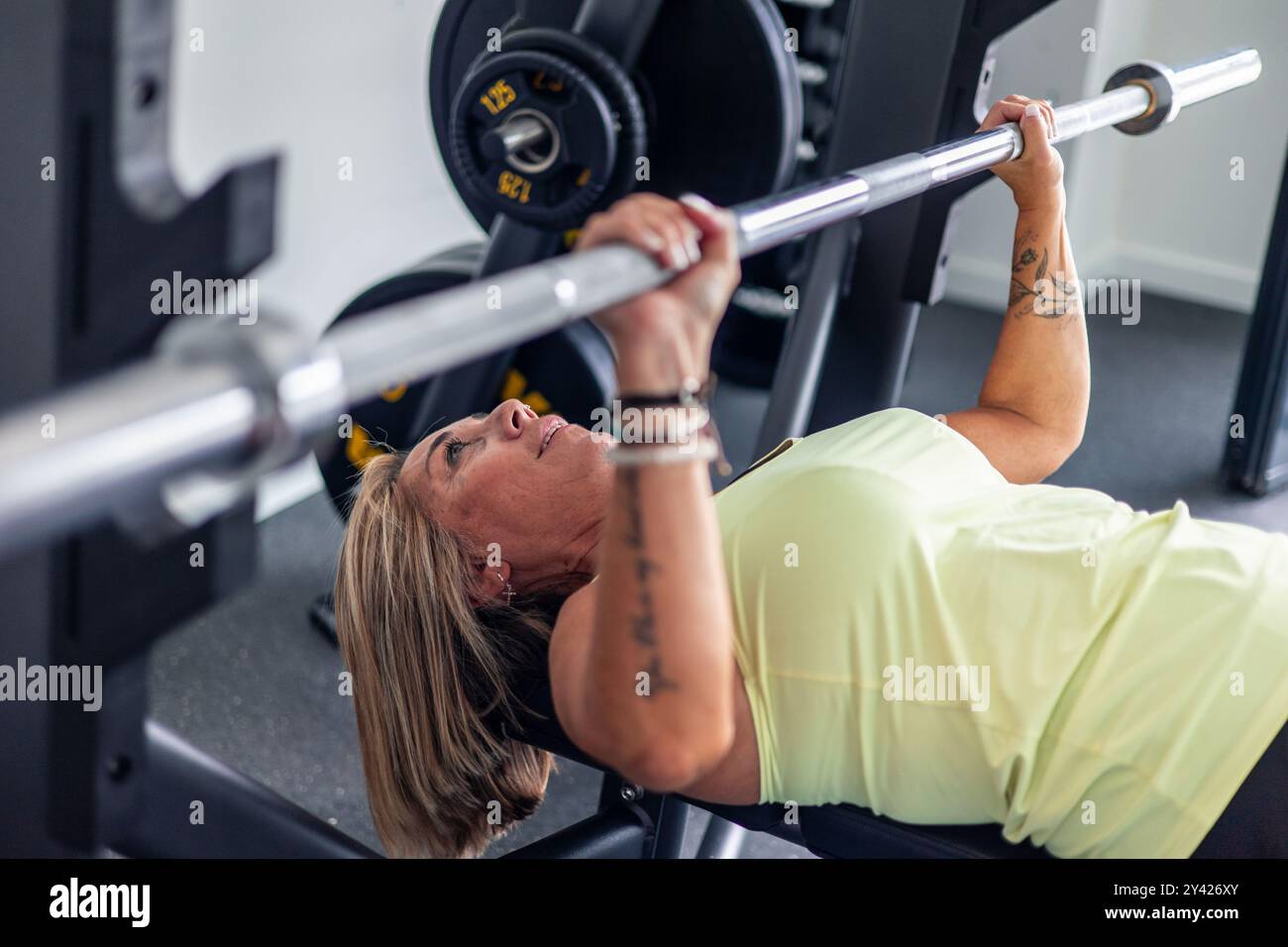 Lächelnde, reife blonde, tätowierte Frau hebt eine Langhantel während einer Bankdruckübung im Fitnessstudio. Das Bild zeigt ihre Stärke und Entschlossenheit Stockfoto