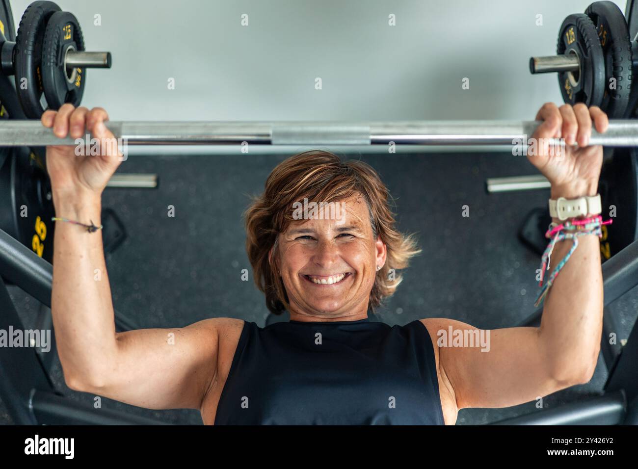 Die Draufsicht zeigt eine ältere Frau, die eine Langhantel hält, während sie während ihres Turntrainings eine Brustdruckübung durchführt. Das Bild erfasst ihren Fokus und ihre Einstellung Stockfoto